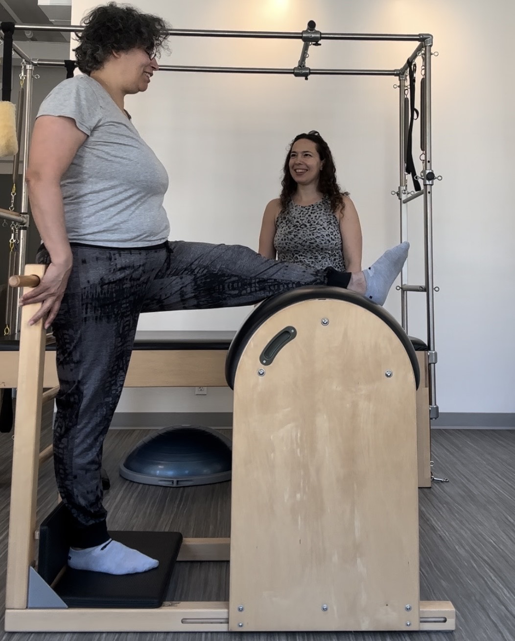 overweight woman over 50 stretching her hamstring on the Pilates ladder barrel as she and her Pilates instructor smile at each other